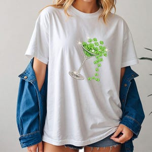 White t-shirt featuring a sparkling martini glass filled with green shamrocks. The glass is embellished with rhinestones, and shamrocks cascade from the glass. A denim jacket is worn over the shirt.