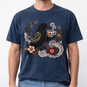 Black crow holding a coffee cup with floral and lace details on a navy Comfort Colors t-shirt