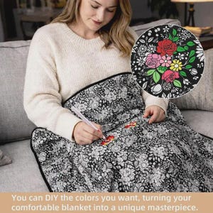 K&ouml;nnte beinhalten: Eine schwarz-wei&szlig; gemusterte Decke mit einer dichten Anordnung von linierten Blumen und Bl&auml;ttern. Ein wei&szlig;er Stift wird verwendet, um bestimmte Bl&uuml;tenbl&auml;tter auf der Stoffoberfl&auml;che mit leuchtenden roten, rosa und gelben Farben auszumalen.