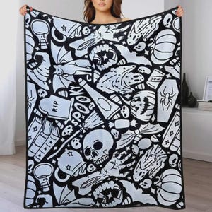 K&ouml;nnte beinhalten: Eine rechteckige Wohndecke mit einem schwarz-wei&szlig;en Muster aus Halloween-Motiven, darunter Totenk&ouml;pfe, K&uuml;rbisse, ein Grabstein mit der Aufschrift RIP, ein Sarg mit einer Spinne, eine Katze, ein Besen und das Wort SPOOKY in gro&szlig;en Buchstaben.