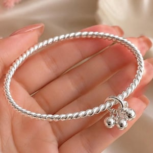 Puede incluir: Una pulsera de tono plateado con diseño de cuerda trenzada y un punto de sujeción circular. Tres pequeñas cuentas esféricas, lisas y de tono plateado cuelgan de una sola anilla, creando un delicado efecto colgante en la banda metálica.