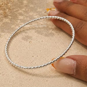 Può includere: Un bracciale rigido circolare e sottile in argento caratterizzato da una texture a corda ritorta continua. La superficie metallica è lucida e riflettente, creando un motivo a spirale uniforme lungo l'intera circonferenza del gioiello.