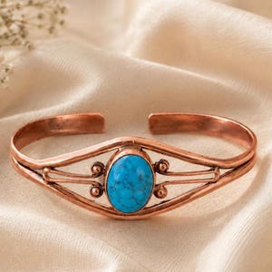 May include: A copper cuff bracelet featuring a central oval-shaped turquoise stone with dark veining. The metal band has a split design that curves around the stone, accented by small spherical metal beads on each side of the central setting.