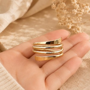 Puede incluir: Un anillo de tono dorado con un diseño de espiral de varias capas. La banda consta de cuatro bobinas distintas, lisas y pulidas, que se envuelven alrededor del dedo, creando una apariencia continua y apilada con un acabado metálico reflectante.