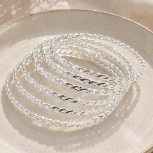 Könnte beinhalten: Ein Set aus sieben silberfarbenen Armreifen, die jeweils eine gedrehte, seilartige Textur aufweisen. Die Armreifen sind übereinander gestapelt und zeigen eine reflektierende metallische Oberfläche sowie eine kreisförmige Form bei allen sieben Stücken.