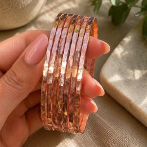 May include: A set of five thin, copper-colored bangles featuring a hammered, textured surface. Each bangle has a reflective, metallic finish with subtle indentations across the exterior, creating a repeating pattern of light and shadow.