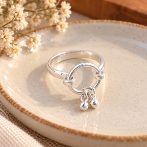 May include: A polished silver ring featuring a circular centerpiece attached to the main band. Two small, round silver beads hang from the bottom of the central circle, creating a dangling effect. The metal has a smooth, reflective finish.