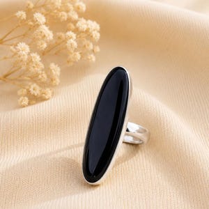 Puede incluir: Un anillo de plata con una piedra negra grande, lisa y de forma ovalada alargada, montada verticalmente. La gema negra pulida está enmarcada por una banda de plata sencilla que se conecta a la parte posterior del engaste, creando un perfil elegante.