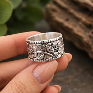 May include: A wide silver-toned band ring featuring an embossed floral pattern with detailed petals and leaves. The edges of the ring are decorated with a raised beaded texture, and the metal has a darkened, oxidised finish in the recessed areas.