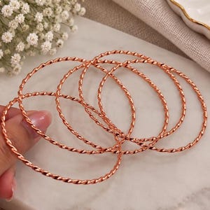 Könnte beinhalten: Ein Set aus vier dünnen, kreisförmigen Armreifen mit einer gedrehten, seilartigen Textur. Die Armreifen bestehen aus einem metallischen Material mit einem warmen, kupferfarbenen Finish. Jeder Armreif weist ein durchgehendes Spiralmuster auf.