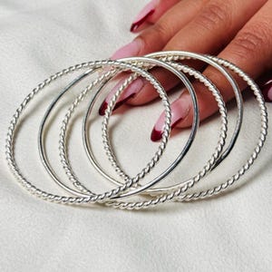 Può includere: Un set di braccialetti d'argento. Alcuni braccialetti presentano un motivo a corda intrecciata, mentre altri sono lisci. I braccialetti circolari sembrano essere in metallo. L'immagine è ripresa da vicino.