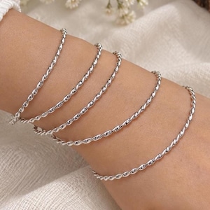 Könnte beinhalten: Ein Set aus vier dünnen, silberfarbenen Metallarmbändern, die jeweils eine gedrehte, seilartige Textur aufweisen. Die Armbänder werden zusammen getragen, umschließen das Handgelenk und haben eine polierte, reflektierende Oberfläche, die das Licht einfängt.