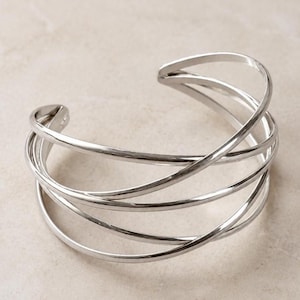 Op de afbeelding: Een zilverkleurige manchetarmband met een meerlaags ontwerp van verschillende dunne, gepolijste metalen banden die elkaar overlappen en kruisen om een open, luchtige structuur te vormen. De armband heeft een glad, reflecterend oppervlak.