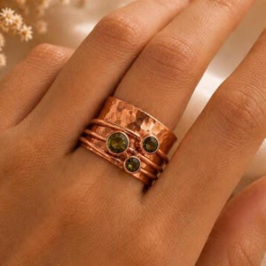 Op de afbeelding: Een brede koperkleurige ring met een gehamerde textuur op de bovenste band en drie gladde, dunne banden eronder. De ring is versierd met drie kleine, ronde, olijfgroene gefacetteerde stenen die afzonderlijk aan de voorzijde zijn gezet.