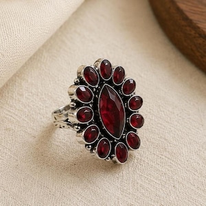 Pode incluir: Um anel de tom prateado com uma grande pedra preciosa central de cor vermelho-escura em corte marquise, rodeada por um halo de doze pedras vermelhas mais pequenas em corte oval. A cravação de metal tem um acabamento texturizado e oxidado.