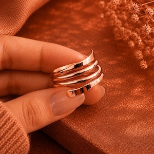 May include: A polished rose gold ring featuring a multi-layered spiral design. The band consists of several smooth, continuous loops that wrap around the finger, creating a stacked appearance with a reflective, metallic finish and curved edges.