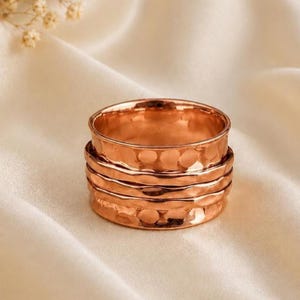 Puede incluir: Un anillo ancho de tono cobre que presenta una superficie martillada y texturizada con múltiples hendiduras horizontales. La banda tiene un acabado pulido y reflectante que capta la luz, creando reflejos sutiles en el metal.