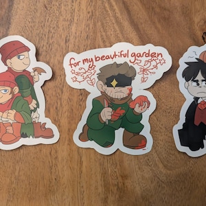 Puede incluir: Tres pegatinas de dibujos animados sobre una superficie de madera. Una pegatina presenta dos figuras con sombreros rojos y trajes verdes. Otra pegatina tiene el texto "for my beautiful garden". La tercera pegatina muestra una figura con un abrigo negro.