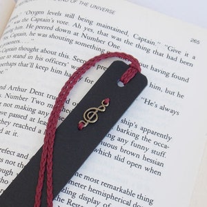 Peut inclure: Un marque-page en cuir noir avec un gland rouge et un charme en forme de note de musique dorée. Le marque-page est ouvert sur une page d'un livre.
