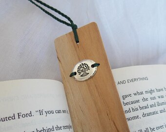 Può includere: Un segnalibro di legno con un cordoncino verde attaccato. Il segnalibro ha un ciondolo d'argento con un disegno di albero. Il segnalibro è appoggiato su un libro con il testo "AND EVERYTHING" visibile.