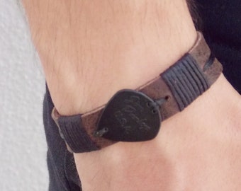May include: A brown leather bracelet with a black guitar pick charm. The bracelet is woven with black thread.