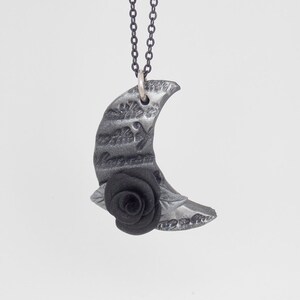 Op de afbeelding: Een zilveren halve maan hanger ketting met een zwarte roos aan de onderkant. De hanger heeft een getextureerd oppervlak met een patroon van schrift.