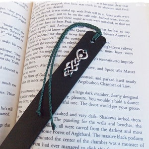 Peut inclure: Un marque-page en cuir marron foncé avec un gland vert et un motif de nœud celtique argenté. Le marque-page est placé sur un livre ouvert avec du texte sur une chambre de droit.
