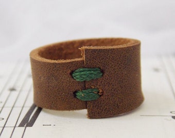 May include: A brown leather cuff bracelet with green stitching. The bracelet is a thick band of textured brown leather, secured with two green stitches. The design is simple and rustic, suitable for everyday wear.