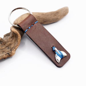 Può includere: Un portachiavi in ​​cuoio marrone con un ciondolo a forma di coda di balena argentata e dettagli in filo blu.