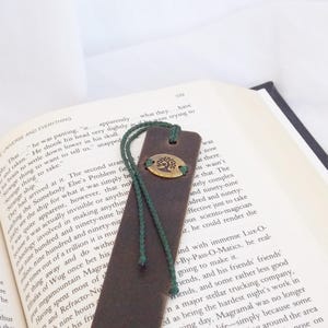 May include: A brown leather bookmark with a green cord and a brass tree charm. The bookmark is lying on a page of a book with text.