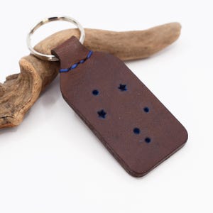 Könnte beinhalten: Brauner Leder-Schlüsselanhänger mit blauem Stepprand und einem ausgeschnittenen Sternbild-Design. Der Schlüsselanhänger hat einen silbernen Schlüsselring.