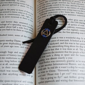 May include: Black leather bookmark with a silver triangle emblem stitched with blue thread. The bookmark has a black cord attached for easy placement in a book. The bookmark is resting on the pages of a book.