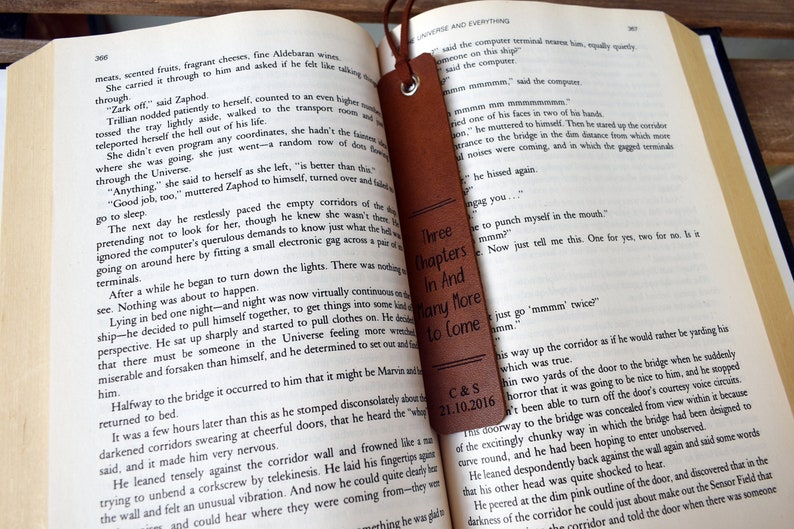 May include: A brown leather bookmark with the text "Three Chapters In And Many More To Come" and the date "C&S 21.10.2016" engraved on it. The bookmark is placed on an open book.
