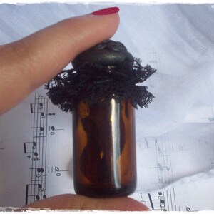 May include: A small, dark brown glass bottle with a black lace trim and a black stopper. The bottle is sitting on a sheet of music.