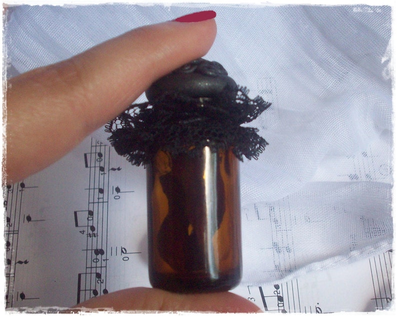 May include: A small, dark brown glass bottle with a black lace trim and a black stopper. The bottle is sitting on a sheet of music.