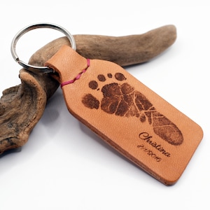 May include: A tan leather keychain with a silver ring. The keychain displays a detailed imprint of a baby's foot, with the name "Christina" and the date "2/11/2016" engraved. The keychain is stitched with pink thread.
