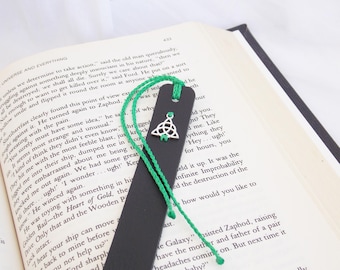 Op de afbeelding: Een zwarte leren bladwijzer met een groene kwast en een zilveren Keltische knoopbedel. De bladwijzer ligt op een boek met tekst.