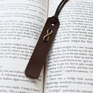 May include: A brown leather bookmark with an infinity symbol charm. The bookmark is laying on an open book with text about a celebrity.