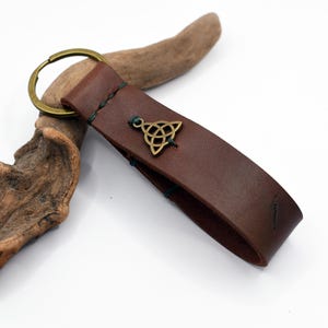 Peut inclure: Un porte-clés en cuir marron avec un anneau de couleur or et un charm en forme de nœud celtique. La lanière en cuir est cousue avec du fil vert. Le porte-clés est présenté sur fond blanc avec un morceau de bois flotté.