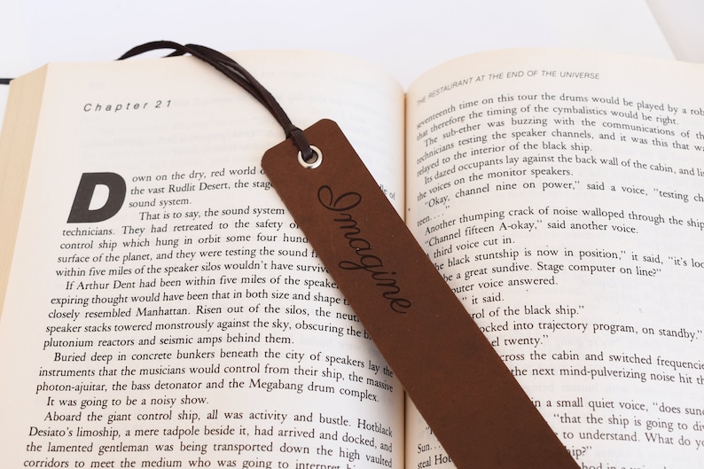 May include: A brown leather bookmark with the word "Imagine" engraved on it. The bookmark is laying on top of an open book with the text "Chapter 21" visible.