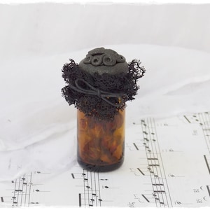 May include: A small amber glass bottle with a black lace and black button embellishment on the lid. The bottle is sitting on a sheet of music.