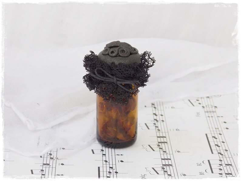 May include: A small amber glass bottle with a black lace and black button embellishment on the lid. The bottle is sitting on a sheet of music.