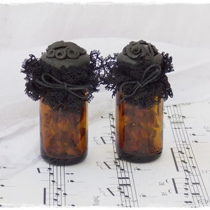 May include: Two small glass bottles with black lace and black clay lids. The bottles are filled with dried flowers and are sitting on a sheet of music.