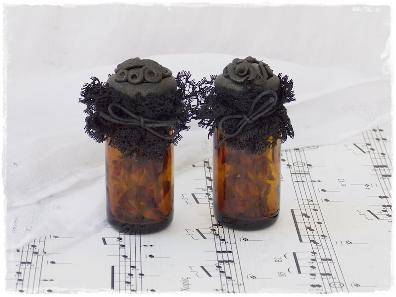 May include: Two small glass bottles with black lace and black clay lids. The bottles are filled with dried flowers and are sitting on a sheet of music.