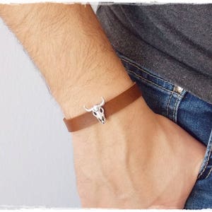 Può includere: Un braccialetto in pelle marrone con un ciondolo a forma di teschio d'argento. Il braccialetto è indossato al polso.