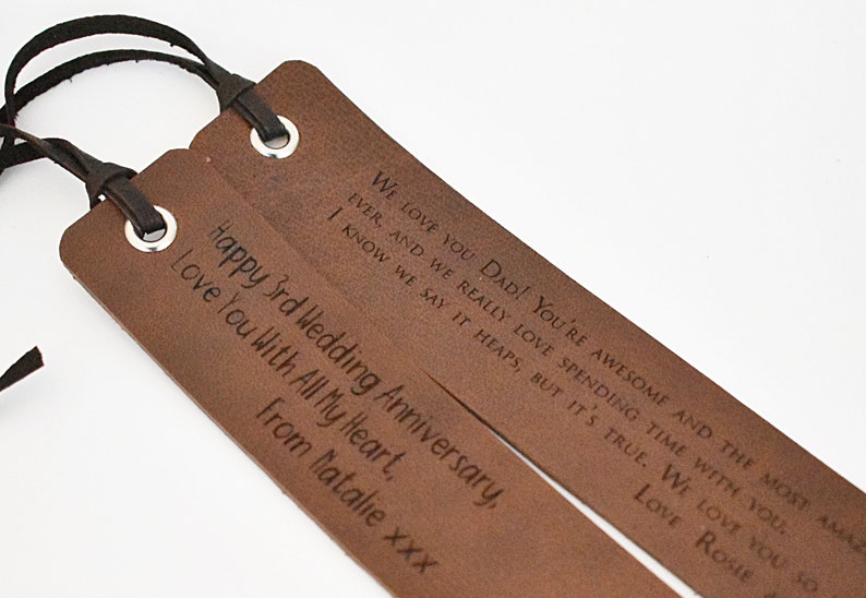 May include: Two brown leather bookmarks with black tassels. The text on the bookmarks reads "We love you Dad! You're awesome and the most amazing ever, and we really love spending time with you. I know we say it heaps, but it's true. We love you so much. Love Rosie" and "Happy 3rd Wedding Anniversary, Love you with all my heart, From Natalie xxx".