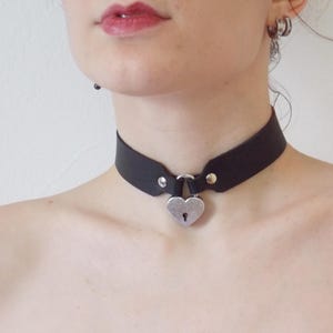 Könnte beinhalten: Ein schwarzes Lederhalsband mit einem silbernen herzförmigen Vorhängeschloss-Anhänger.