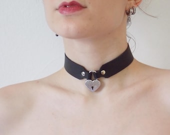 Könnte beinhalten: Ein schwarzes Lederhalsband mit einem silbernen herzförmigen Vorhängeschloss-Anhänger.