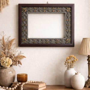 May include: A rectangular, dark brown picture frame with ornate detailing in gold and silver tones, hanging on a white wall. The frame is empty, with a decorative border. The image suggests home decor and wall art.