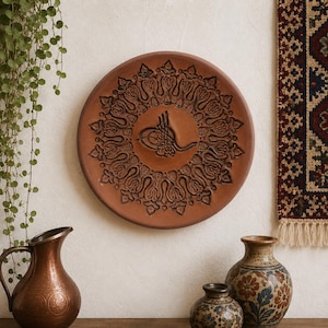 Puede incluir: Un plato decorativo redondo, de color terracota, con un diseño floral en relieve y un emblema central. El plato se exhibe en una pared, con una jarra de cobre y dos jarrones de cerámica en primer plano.